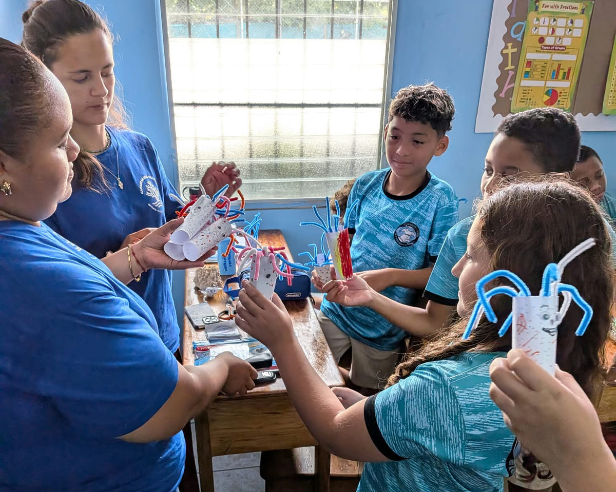 A group of children and two adult women gathered around a table in a classroom, holding and assembling small white and blue coral polyp models. The room has light blue walls, a tiled floor, and a window in the background.