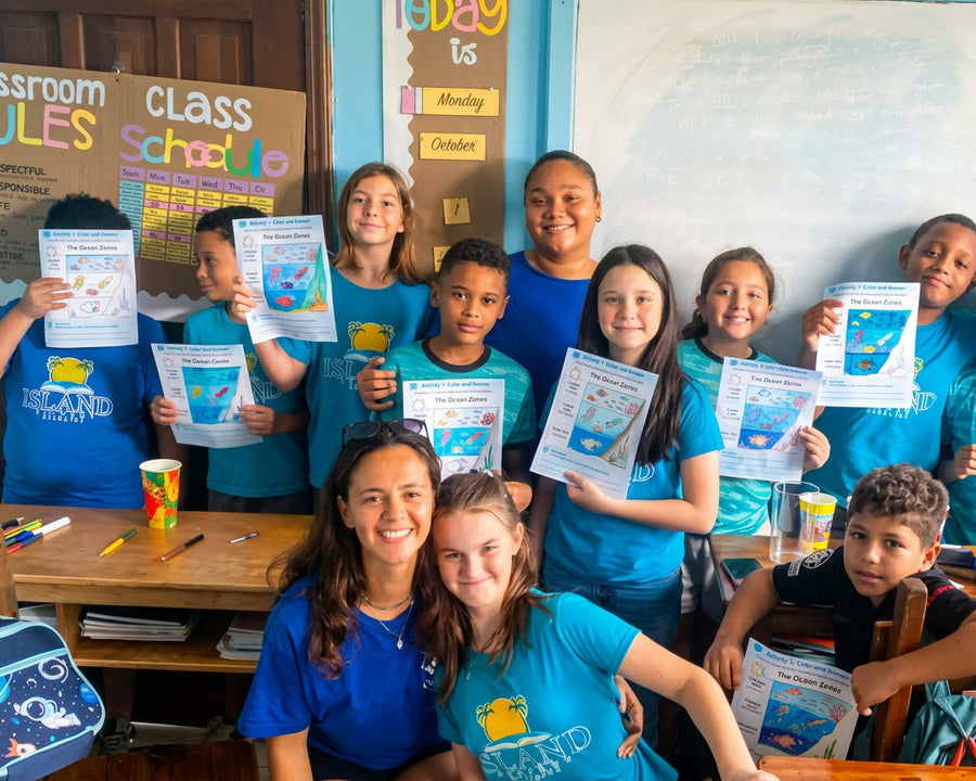 A group of students and an adult teacher wearing matching blue t-shirts with a 'Classroom Buddies' logo, holding up printed Ocean worksheets in a classroom setting.
